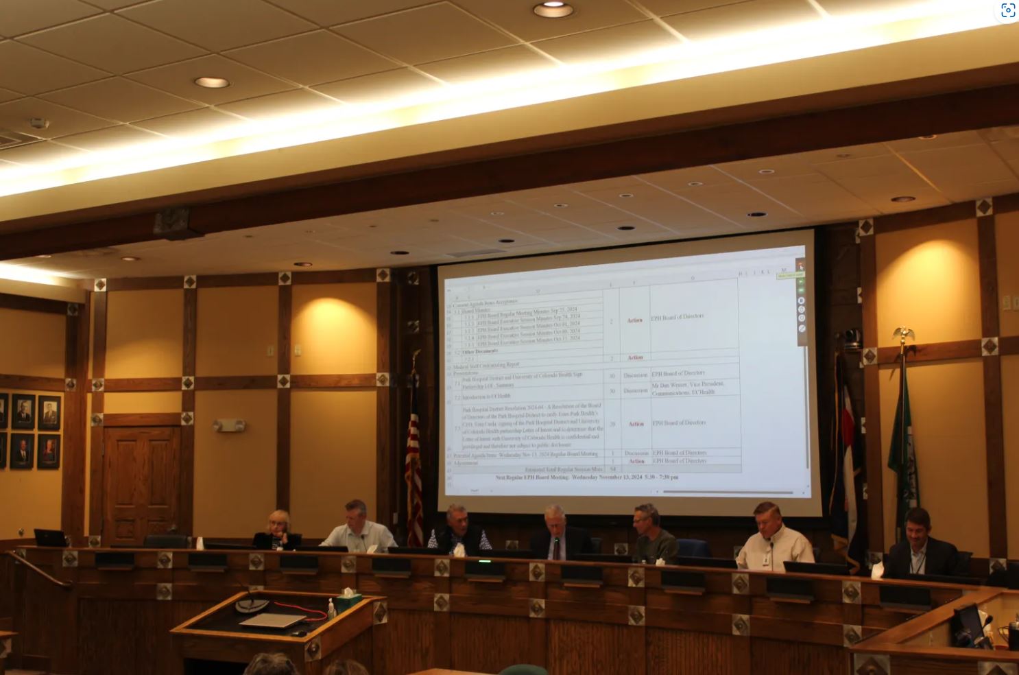 A city council meeting in session with seven officials seated at a long table, a large screen displaying agenda items, and flags visible in the background.