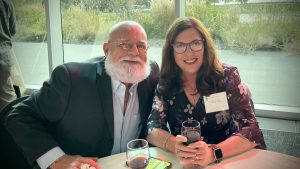 A man and a woman sit at a table with glasses of red wine. They are smiling and have name badges. The table is in front of a window with greenery outside.