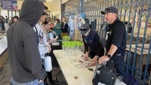 People at a table display with plastic bottles and electronic equipment, engaging in conversation. Indoor setting with a metal fence in the background.