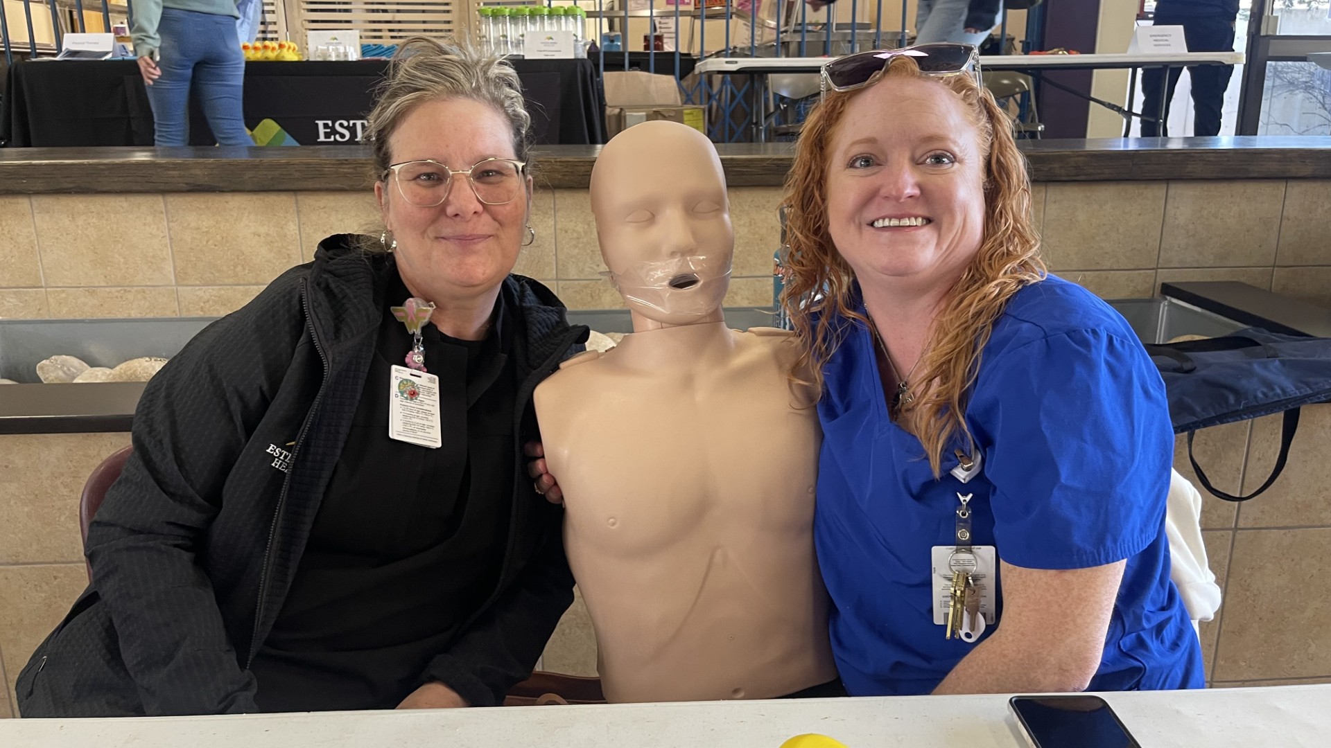 Two women are seated on either side of a CPR training mannequin, smiling at the camera. The women are wearing ID badges and casual clothing.