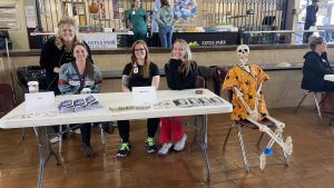 Four women and a dressed skeleton sit at a table with informational materials inside a community center.