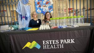 Two people at an Estes Park Health booth with promotional items, including safety gear, hand sanitizers, and cups.