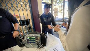 A person demonstrates a medical device, showing how it works, with another person observing. There is equipment on the table, and a sign reads "Emergency Response.