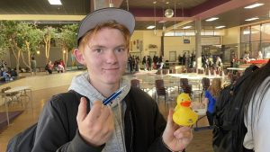 A person stands indoors in a busy area, holding a pen and a yellow rubber duck, smiling slightly.