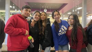 Five people stand closely together and smile. Three wear hoodies, one wears a blue adidas hoodie, another wears a burgundy shirt. They are in an indoor public area with pendent lighting.