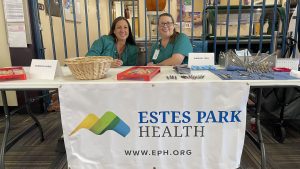 Two women in medical uniforms sit at a table with surgical tools and a sign reading "Estes Park Health" at a health event.