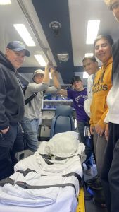 A group of five people stand inside an emergency vehicle next to a stretcher. Some are smiling and wearing casual clothing.