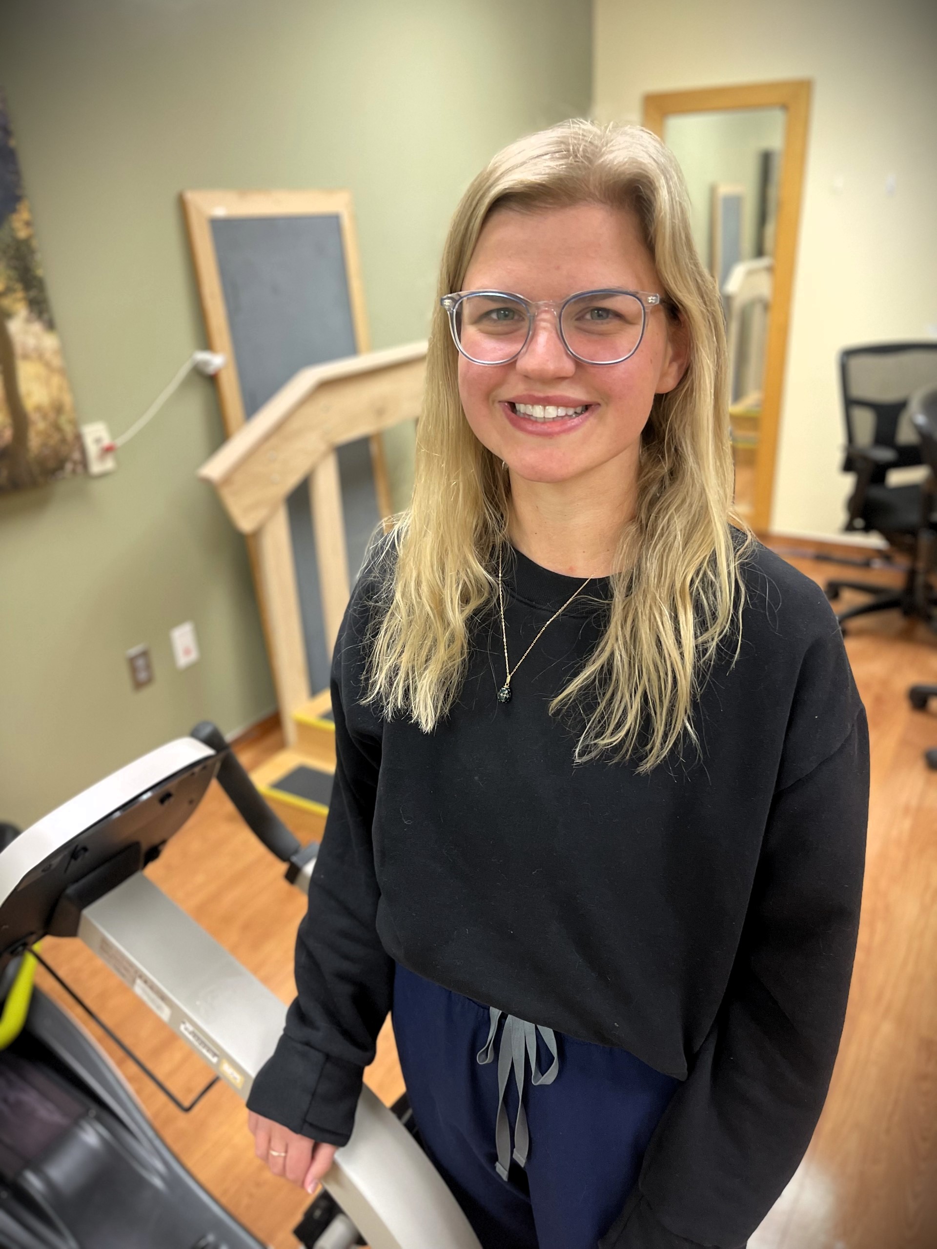 A person with long hair and glasses stands in a room with office furniture and equipment, smiling at the camera.