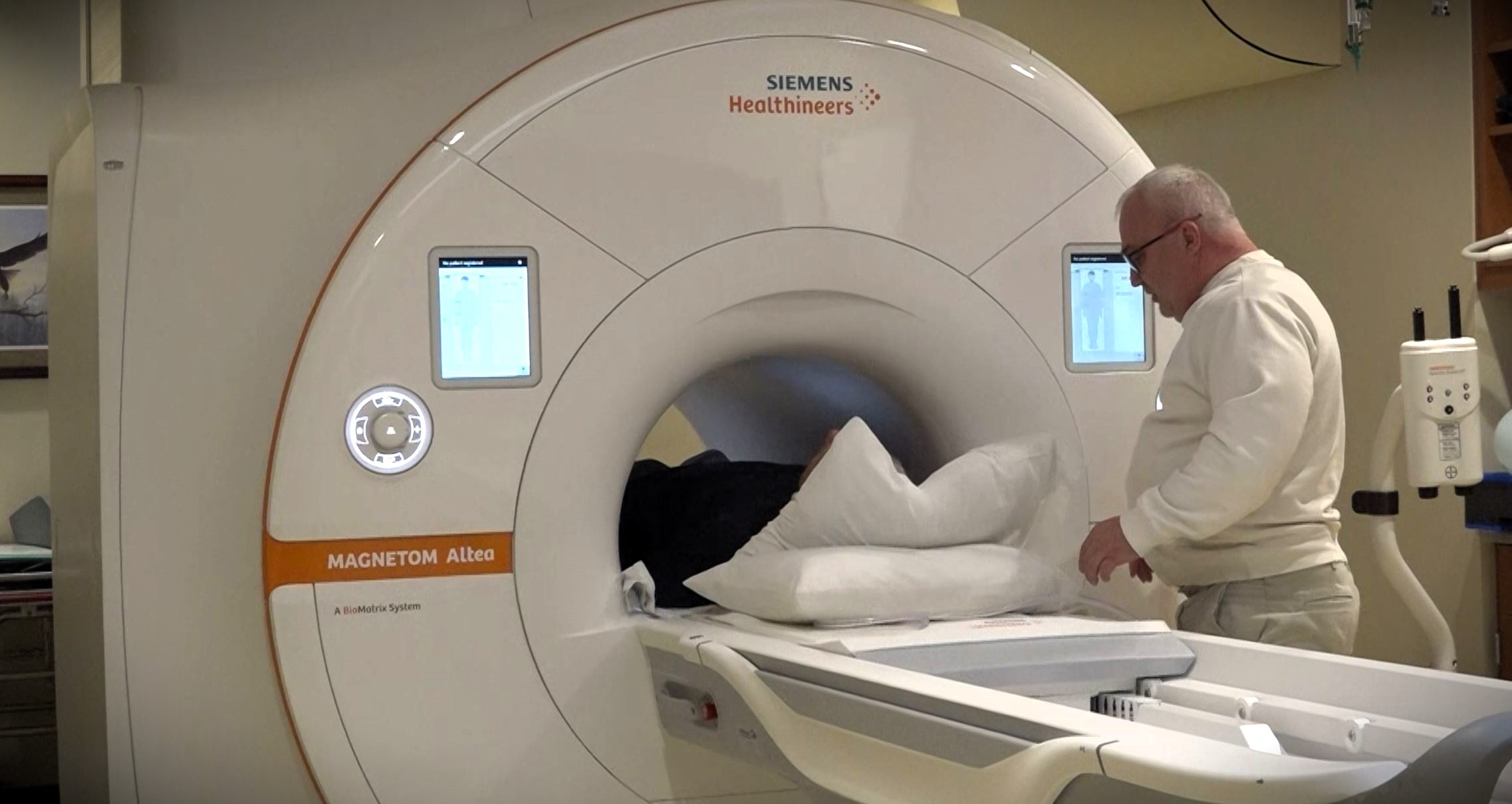 A person prepares to enter a Siemens MAGNETOM Altea MRI machine, assisted by a man in a white sweater.