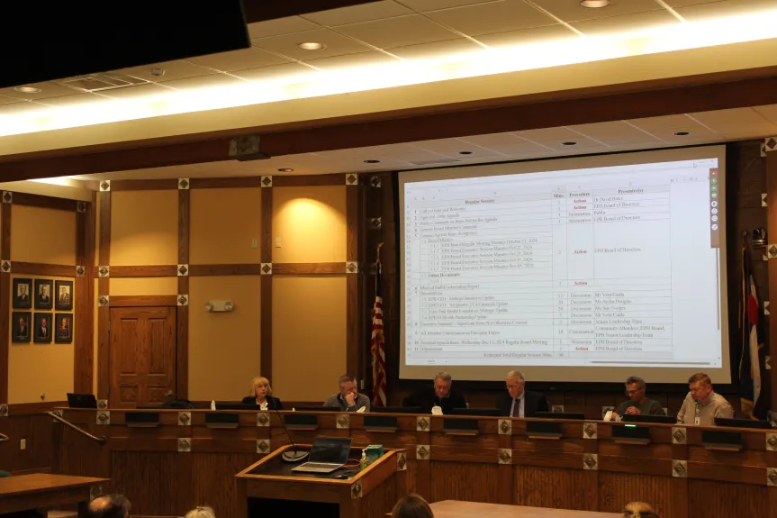 A council meeting in session with six people seated at a long desk, a large screen displaying text, and an American flag in the background.