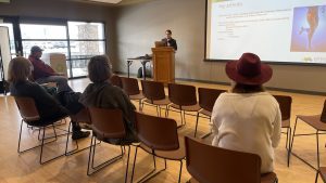 A person stands at a podium giving a presentation on hip arthritis to a small audience in a conference room.