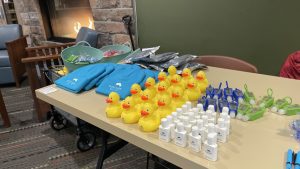 Table with rubber ducks, hand sanitizers, blue hats, sealed bags, and green carabiners in front of a fireplace.