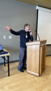 A person speaks into a microphone while gesturing with one hand, standing at a wooden podium in a room with a projection screen.