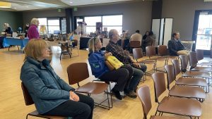 People sitting on chairs in a community center with book stalls and tables in the background.