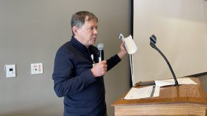 Person holding a folded piece of paper and speaking into a microphone at a podium with a stand.