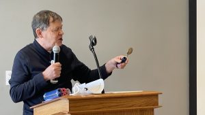 A person holding a microphone in one hand and a spoon in the other, standing at a podium with papers and speaking equipment.