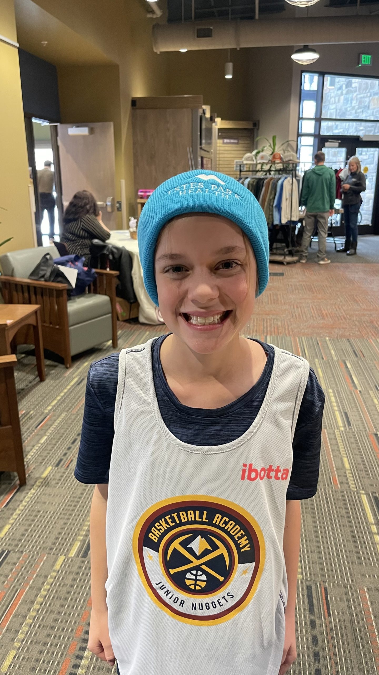 A smiling child wearing a blue beanie and a basketball-themed jersey stands indoors. People and furniture are visible in the background.