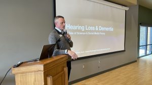 A man gives a presentation on "Hearing Loss & Dementia" with a podium and screen in the background.