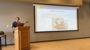 A person presents a lecture on hip pain at a podium, with a slide showing information and an image of a patient on crutches.