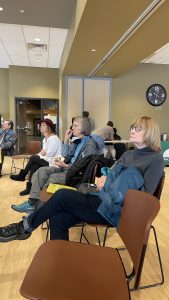 A group of people sitting attentively in a room with beige walls, some taking notes. A clock reads 12:10.