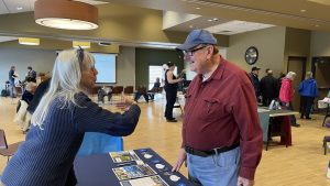 Two people talking at an indoor event with informational tables. One person is gesturing animatedly while the other listens and smiles.