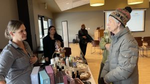 Two women at a vendor table with various skincare products. One woman is wearing a coat and hat, while the other stands behind the table. Other people are in the background.