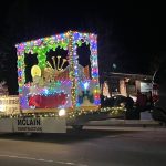 A parade float features a colorful, illuminated display with musical instruments, labeled "Christmas Calliope," towed by a vehicle adorned with lights.