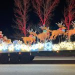 A festive parade float featuring Santa Claus in a sleigh pulled by reindeer, surrounded by bright lights and illuminated snowflake decorations, moves along a street at night.