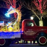 A red truck decorated with Christmas lights, featuring a light-up giraffe and small houses, drives in a nighttime parade with illuminated trees in the background.