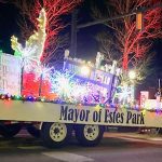 A holiday parade float labeled "Mayor of Estes Park" is adorned with lights, presents, and a reindeer figure, set against a backdrop of bare trees illuminated in red.