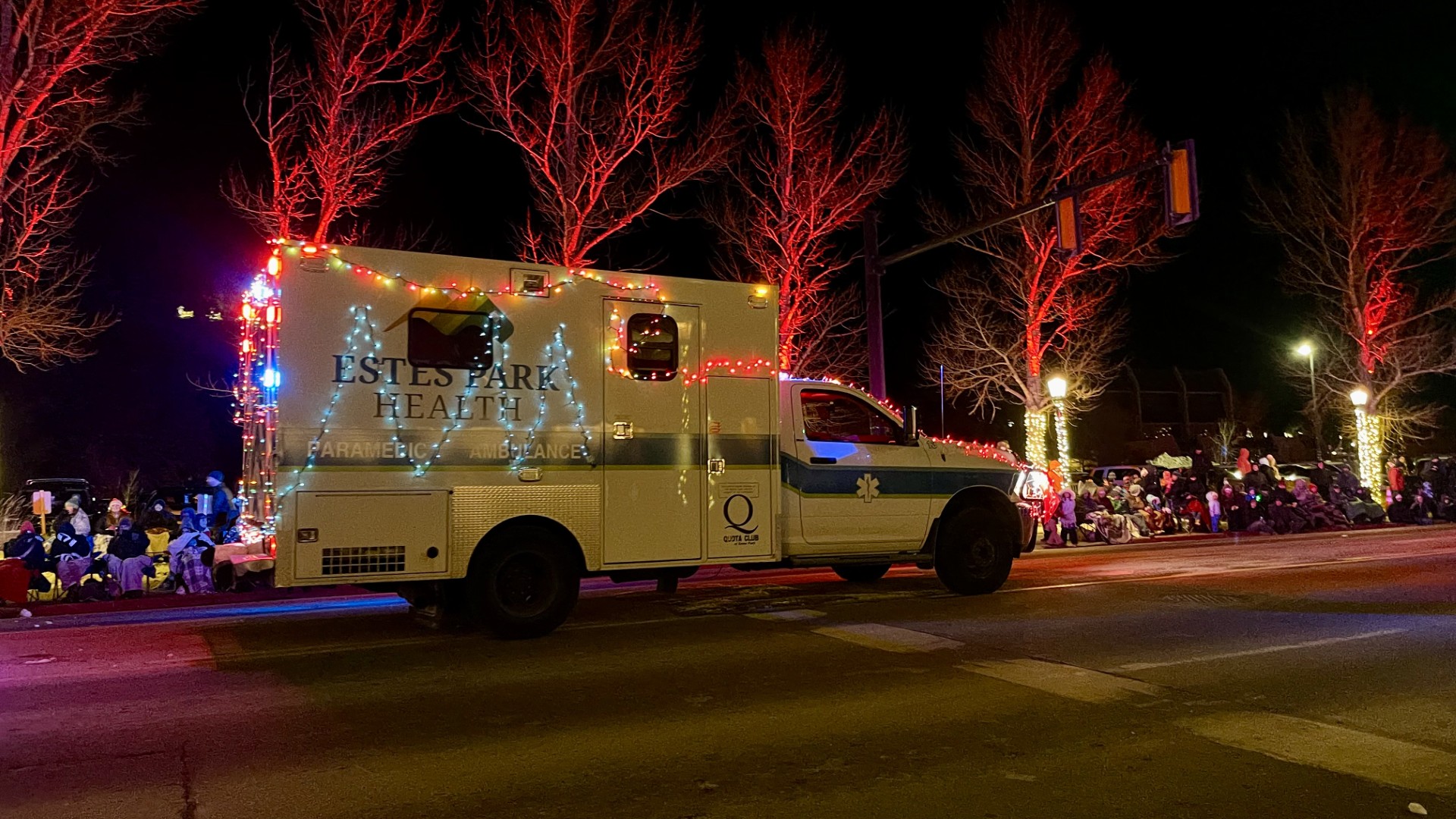 Ambulance decorated with festive lights drives down a street at night, lined with spectators and illuminated trees.