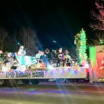 A holiday parade float labeled "EHS Holiday Band" travels at night, decorated with colorful lights and carrying people with musical instruments.