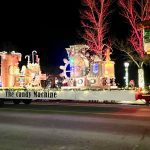 A brightly lit parade float named "The Candy Machine" moves down a street at night, displaying colorful decorations and lights.