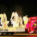 A parade float titled "The Angels Sing" with illuminated angel figures is pulled by a truck at night.