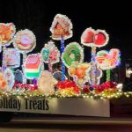 A festive parade float features illuminated holiday-themed decorations, including cookies and candy, labeled "Holiday Treats.