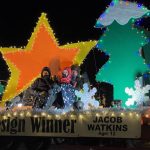 Parade float with holiday decorations, lit star and trees, winning design by Jacob Watkins, age 12, featuring three people, two adults and one child, standing on it.