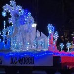 A parade float decorated with blue and white lights features the words "Ice Queen" and ice-themed embellishments, moving along a street at night.