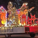 A festive parade float decorated with Christmas lights, featuring a musician playing a guitar and holiday-themed decorations like stars and artificial trees.