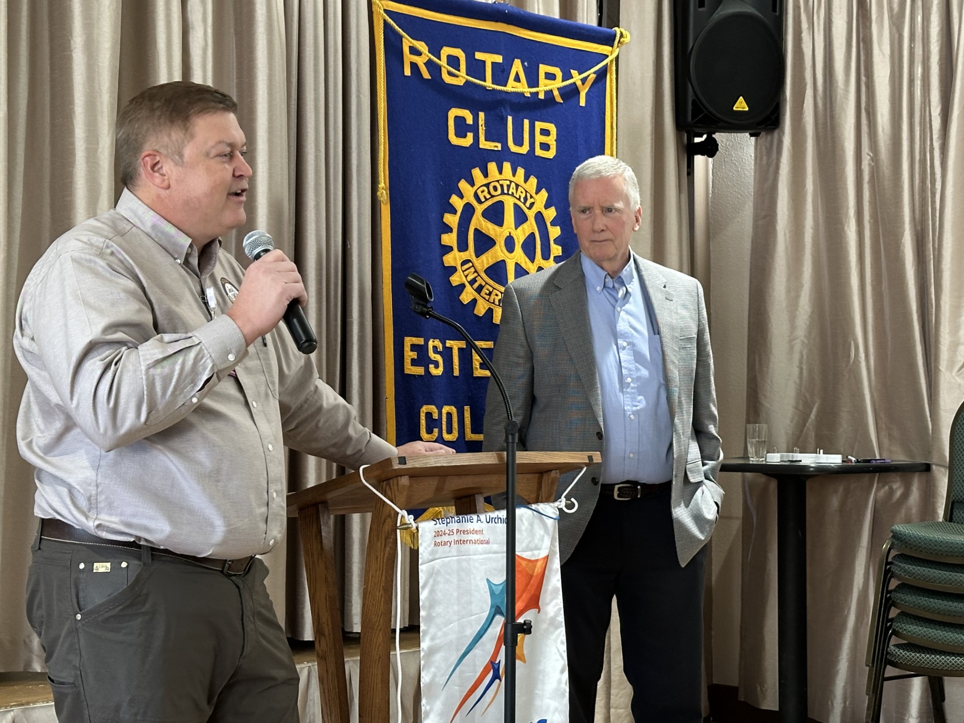 Two men stand at a podium in front of a Rotary Club banner, one speaking into a microphone while the other listens.