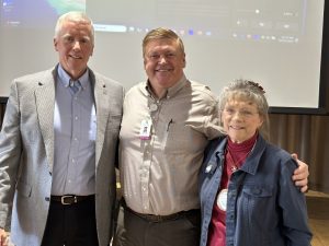 Three people smiling and standing in front of a screen. The two men are wearing business attire, and the woman is wearing a denim jacket with a pin.