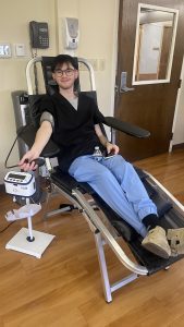 A person in medical scrubs donates blood while seated in a reclining chair, using a machine to monitor the process in a clinical setting.