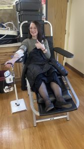 A woman sitting in a chair giving a thumbs up while donating blood, with a blood pressure cuff on her arm.