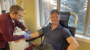 A person in a chair donates blood while a healthcare worker assists. The room has large windows with a view of trees outside.