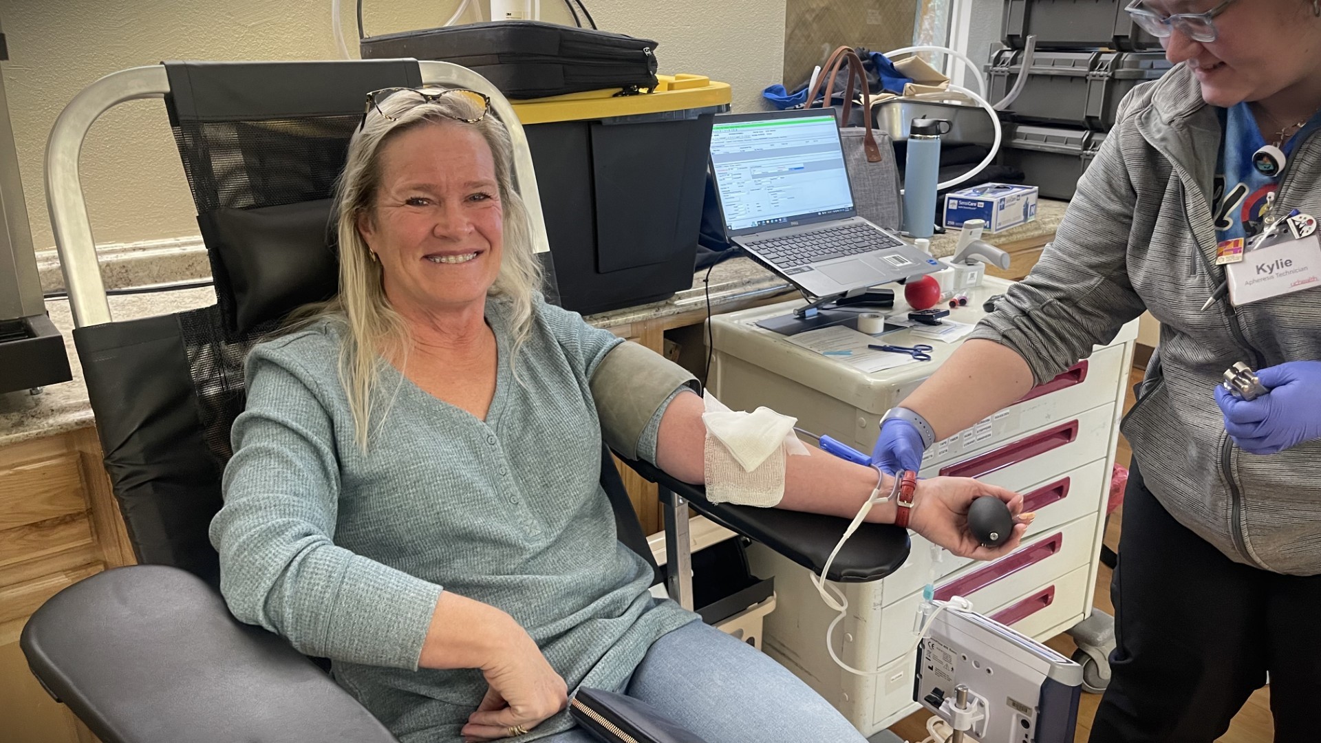 A woman donates blood while sitting in a chair. A healthcare worker assists her, and medical equipment is visible in the background.