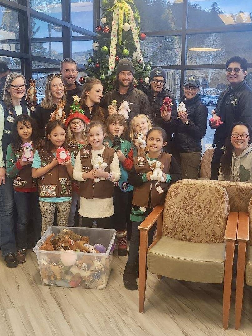 A group of adults and Girl Scouts pose indoors with stuffed animals in front of a decorated Christmas tree and large window.