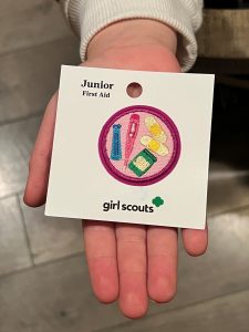 A person holds a Girl Scouts "Junior First Aid" badge featuring a needle, band-aid, and first aid supplies on a card.