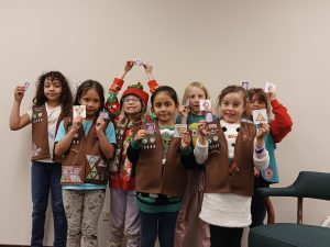 A group of seven young girls in brown vests proudly display various badges in a room. Some are smiling, and others are raising their badges in the air.