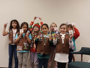 A group of young girls in brown vests proudly display badges they earned. They are standing indoors against a plain wall.