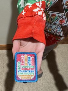 A person wearing a festive sweater holds a patch that says, "FEED, HELP, BUILD, SUPPORT, DONATE, VOLUNTEER, GIRL SCOUTS, COMMUNITY SERVICE.