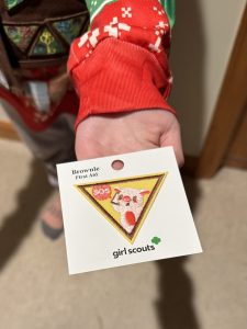 A person holds a Girl Scouts Brownie First Aid patch with an image of a cat wearing a bandage, labeled "SOS.